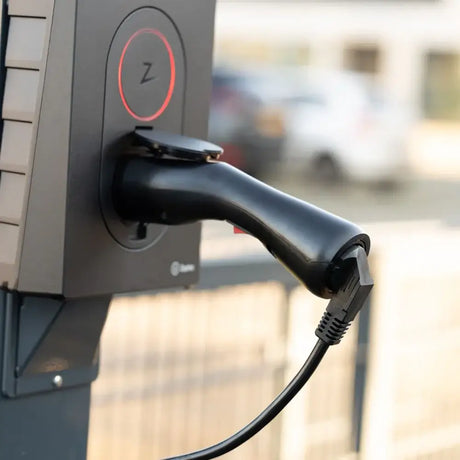 Close-up van een Adapter Type 2 (laadstation) naar Type F 230V schuko-stopcontact met een zwarte oplaadstekker aangesloten. De achtergrond is onscherp, waardoor er een hint van buitenelementen en een hek te zien is.