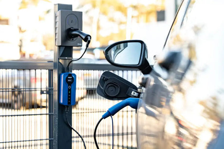 Een close-up van een Adapter Type 2 (laadstation) naar Type F 230V schuko-stopcontact aangesloten met een blauwe laadkabel aan een elektrisch voertuig, buiten getoond met een hek op de achtergrond.