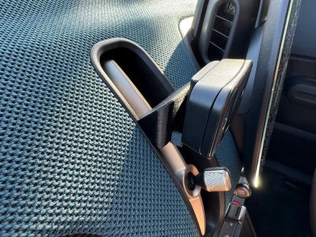 Een close-up van de Compacte Flitsmeister TWO houder voor de MINI Countryman U25 op een ventilatieklep op het dashboard, met links de gestructureerde stoelbekleding en rechts een deel van het dashboard en de ventilatiebediening van de MINI Countryman U25.