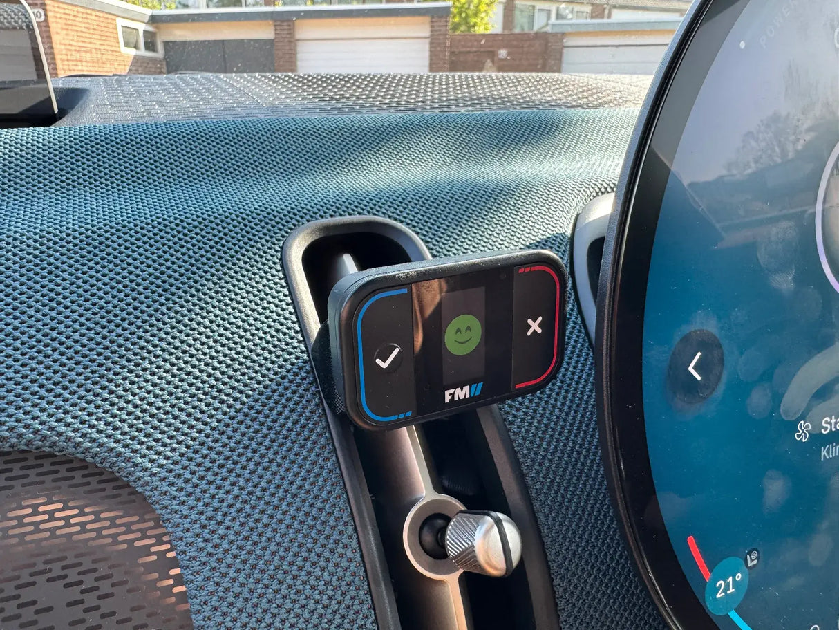 Een close-up toont de Compacte Flitsmeister TWO houder voor de MINI Countryman U25 met een groen lachend gezichtje, vinkje en X bevestigd aan een ventilatierooster bij het ronde digitale display. Garages en een zonnige hemel verschijnen op de achtergrond.