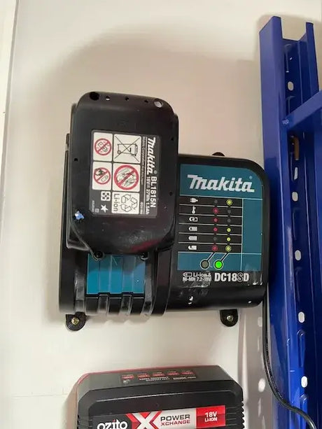 Een Makita 18V DC18RD Lader Wandmontage Voet houdt de lader stevig vast aan de muur, met de accu bovenaan bevestigd. Indicatielampjes en labels zijn zichtbaar en een blauw metalen rek verschijnt gedeeltelijk rechts.