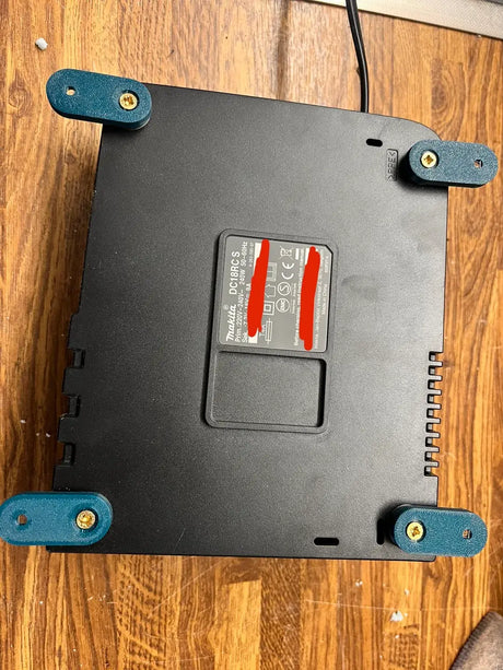 Een Makita 18V DC18RD Lader Wandmontage Voet, zwart met vier groenblauwe voetjes bevestigd met goudkleurige schroeven, ligt ondersteboven op een houten oppervlak. Het label wordt gedeeltelijk verborgen door rode markeringen.