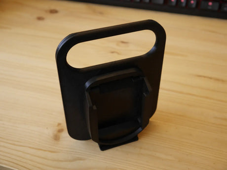 Een Makita LXT 18V BL1830-BL1890 Accuhouder & Transport Drager | 3D Geprint, zwart plastic met een rechthoekig handvat en schuine houder, staat op een licht houten bureau met een toetsenbord op de achtergrond.