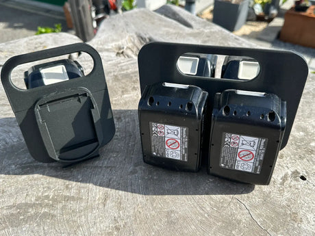 Twee zwarte accu's met veiligheidslabels liggen buiten op een houten oppervlak, naast een rechtopstaande Makita LXT 18V BL1830-BL1890 Accuhouder & Transportwagen | 3D Geprint. Zonlicht werpt scherpe schaduwen.