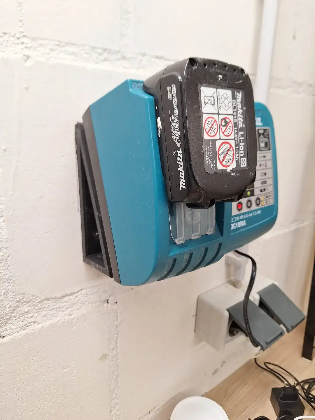 Een Makita Lader Wandhouder DC18RA met een accu is gemonteerd op een witte bakstenen muur boven een stopcontact en stekkerdoos in een werkplaats.