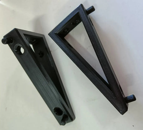 Twee zwarte, driehoekige Makita oplader Wandhouder DC18RA beugels met gaten en haringen zijn 3D-geprinte plastic bevestigingen voor Makita oplader wandmontage, afgebeeld schuin op een wit oppervlak.