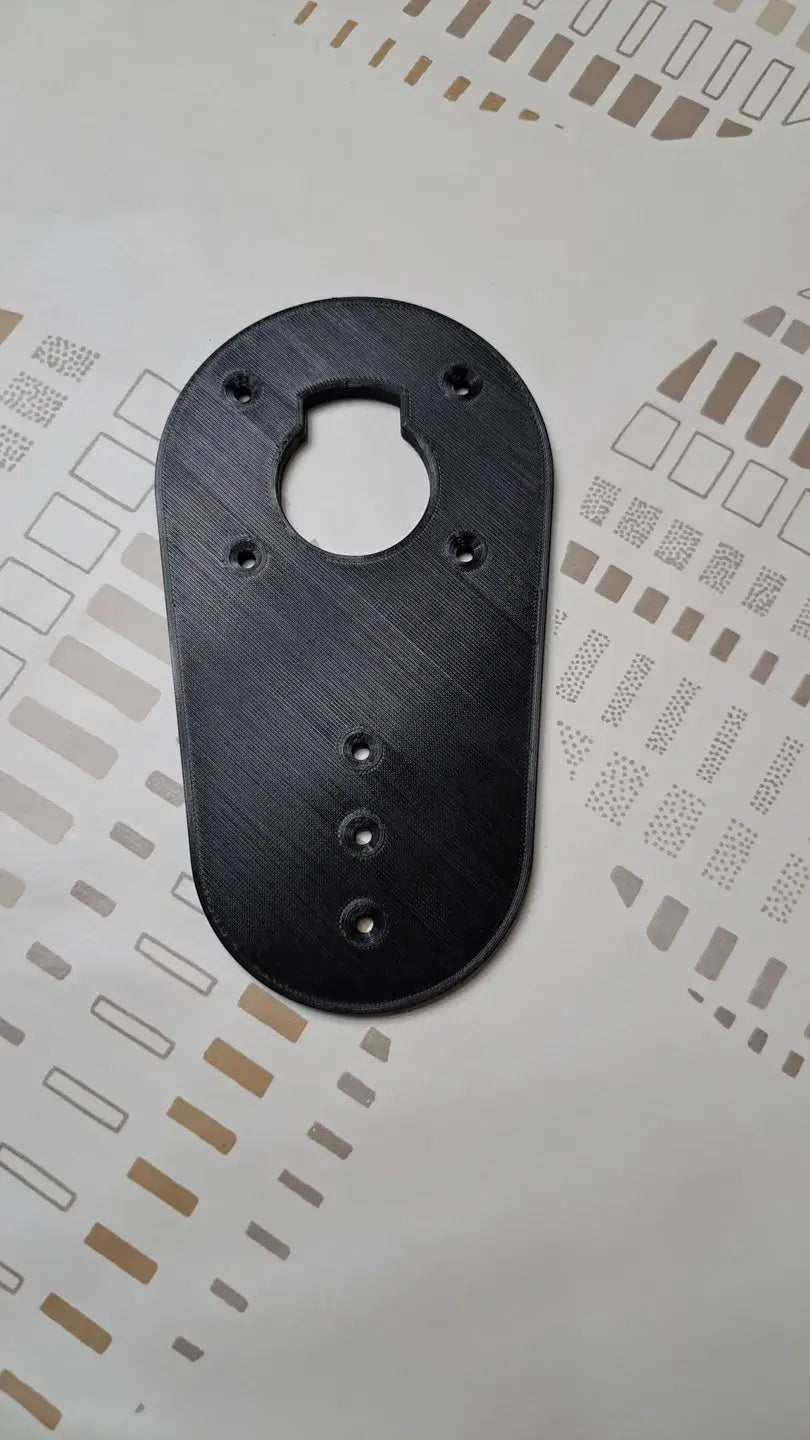 Een Makita RT0700 Bovenfrees Basisplaat, zwart en ovaalvormig met meerdere gaatjes, rust op een wit oppervlak met bruine en bruine geometrische patronen.