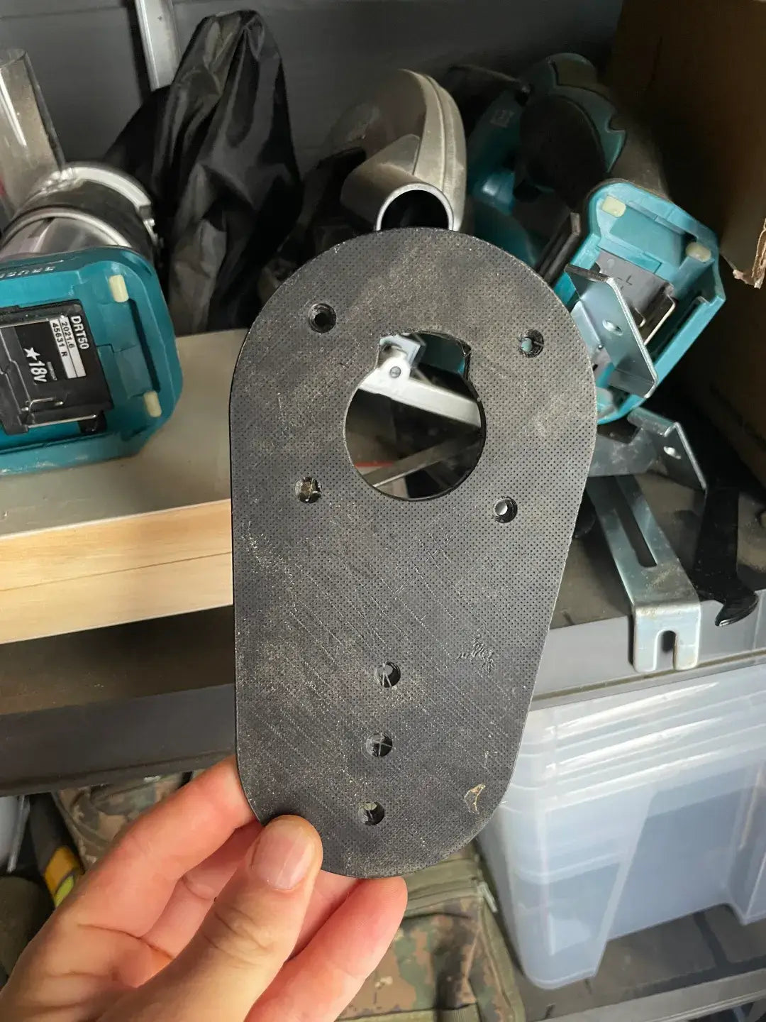 Een hand houdt een Makita RT0700 Bovenfrees Basisplaat vast, een zwarte basisplaat voor bovenfrezen, in een werkplaats met gereedschap en opbergdozen zichtbaar op de achtergrond.