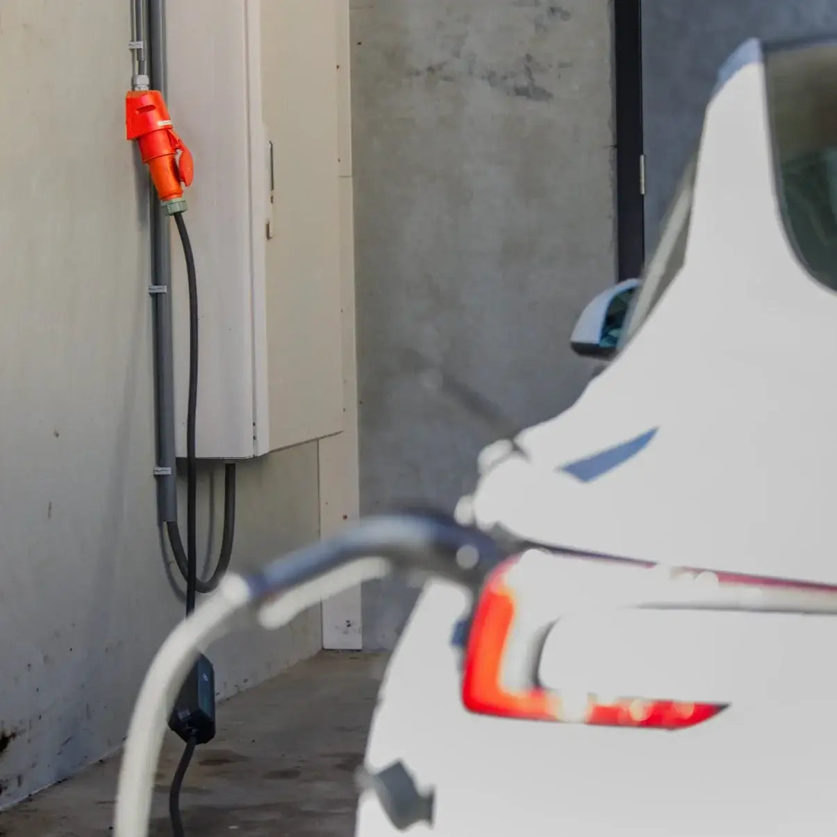 Een witte elektrische auto wordt opgeladen op een betonnen parkeerplaats met behulp van de 🇳🇱 Type 2 CEE 32A 3-Fase Mobiele Laadkabel Verstelbaar 22kW, aangesloten op de achterkant van de auto. Het product heeft 3 jaar garantie en is gemaakt in Nederland.