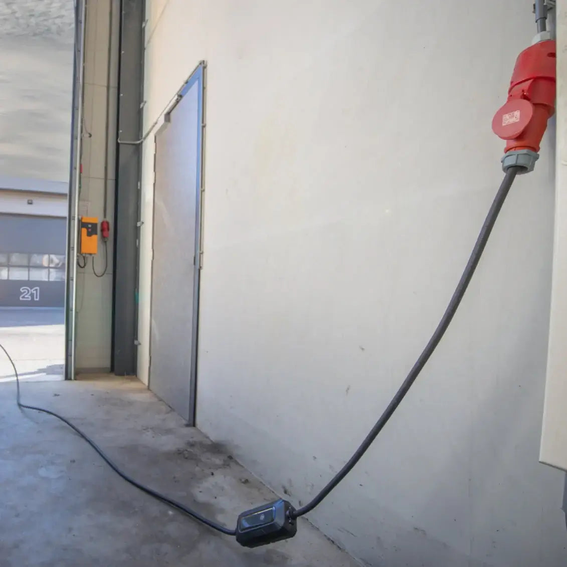Een 🇳🇱 Type 2 CEE 32A 3-Fase Mobiele Laadkabel Verstelbaar 22kW (Nederlands product, 3 jaar garantie) is aangesloten op een rood stopcontact, dat langs een betonnen vloer loopt in een industrieel gebouw in de buurt van een grijze metalen deur en elektriciteitskast.