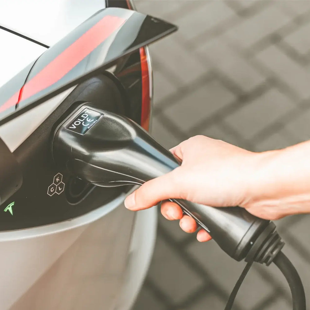 Een hand steekt een laadkabel van de 🇳🇱 VOLDT Booster Master Travelset - 22kW 3-Fase Professionele Laadoplossing in een witte elektrische auto, om deze slimme mobiele EV lader te demonstreren op een verharde ondergrond.