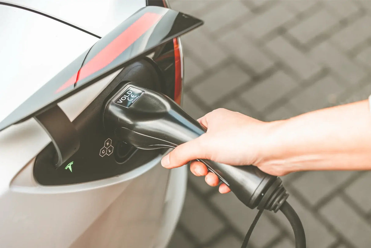 Een hand steekt een laadkabel van de 🇳🇱 VOLDT Booster Master Travelset - 22kW 3-Fase Professionele Laadoplossing in een witte elektrische auto, om deze slimme mobiele EV lader te demonstreren op een verharde ondergrond.