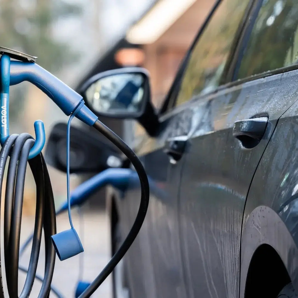 Een VOLDT Type 2 Mode 3 16A 3-Fase Laadkabel (11kW, 3 jaar garantie, Nederlands product) sluit een elektrisch voertuig aan op een laadstation buiten in een woonwijk, met vage bomen en gebouwen op de achtergrond.