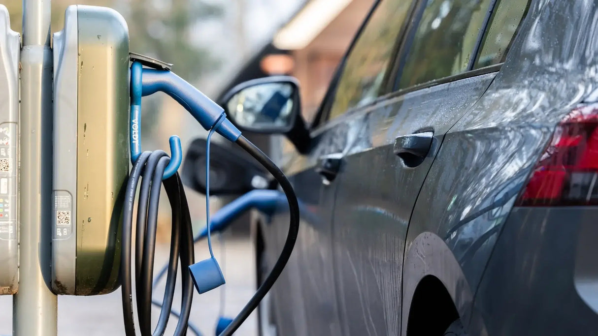 Een VOLDT Type 2 Mode 3 16A 3-Fase Laadkabel (11kW, 3 jaar garantie, Nederlands product) sluit een elektrisch voertuig aan op een laadstation buiten in een woonwijk, met vage bomen en gebouwen op de achtergrond.