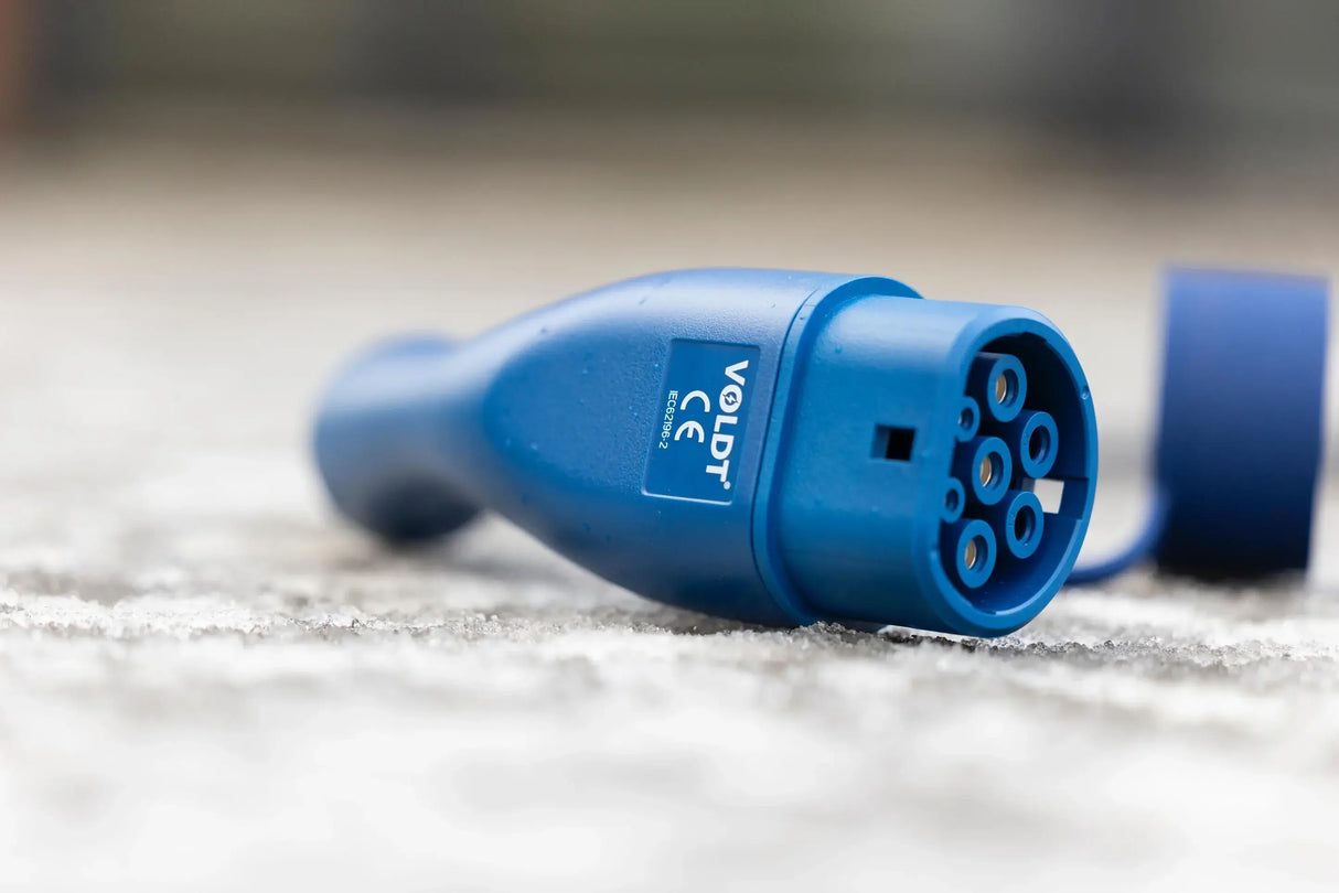 Een close-up van de 🇳🇱 VOLDT Type 2 Mode 3 16A 3-Fase Laadkabel 11kW, een in Nederland geproduceerde laadkabel voor EV's met een rond uiteinde met meerdere pinnen en zichtbare branding, ligt op beton. Inclusief 3 jaar garantie.