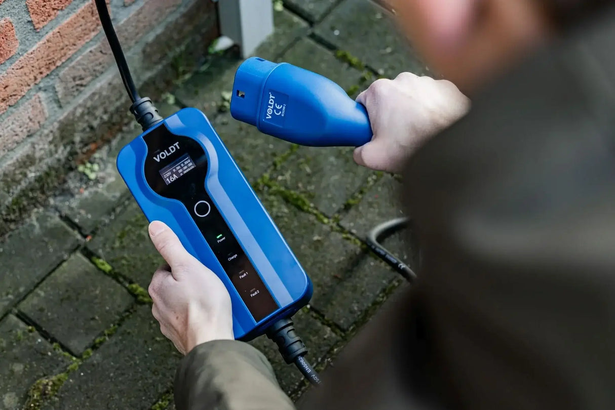 Een persoon houdt een Voldt Type 2 CEE 16A Mobiele Laadkabel (1-Fase, 3,7kW) vast, klaar om zijn elektrische auto buiten op te laden op een verharde ondergrond in de buurt van een bakstenen muur.