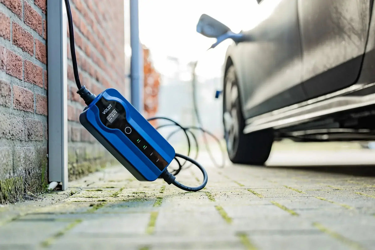 Een close-up van een zwarte elektrische auto-oplader met de Voldt Type 2 CEE 16A Mobiele Laadkabel 1-fase instelbaar 3,7kW (Nederlands product, 3 jaar garantie) bij een bakstenen muur, met het apparaat en de snoeren zichtbaar op de grond.