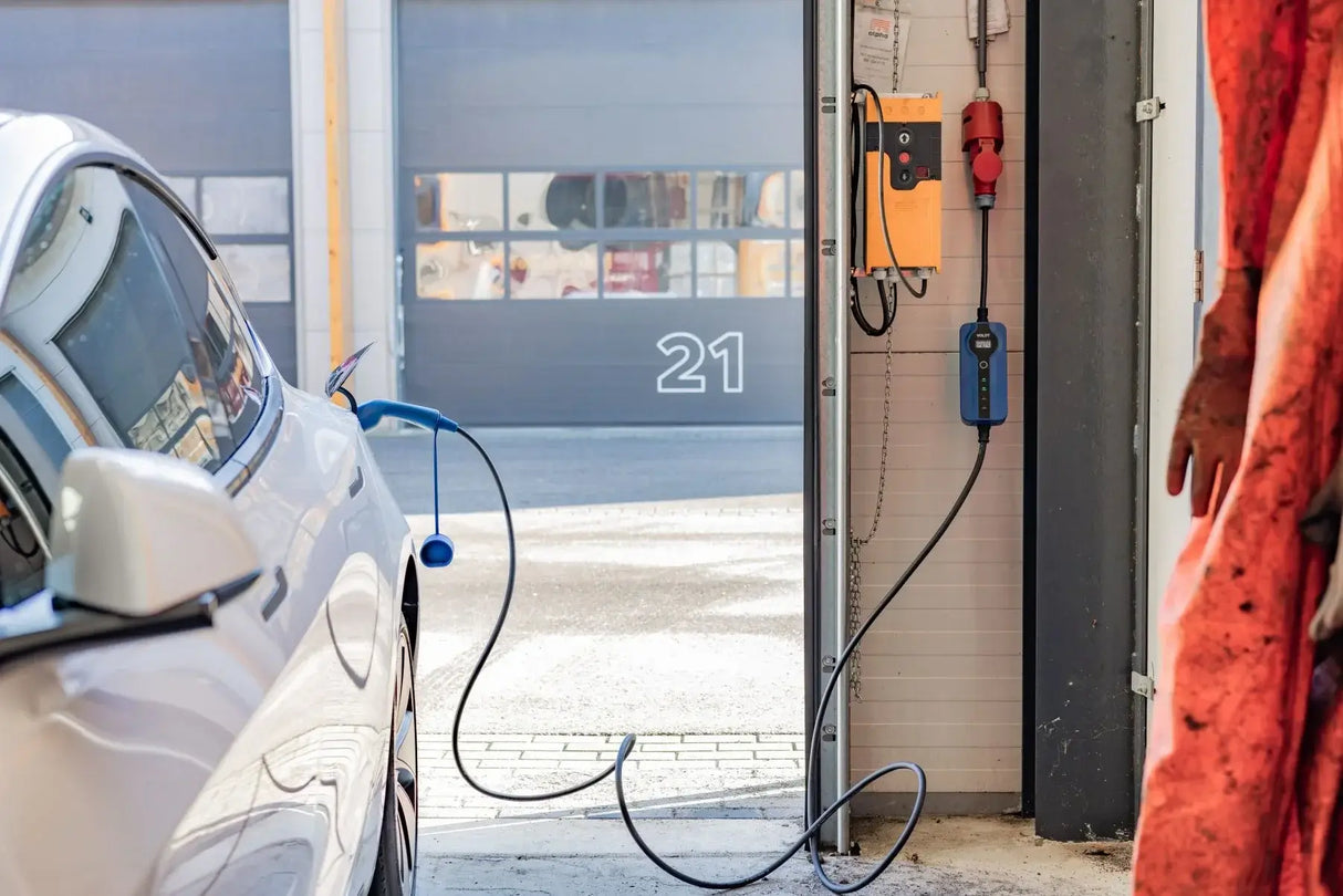 Een witte elektrische auto staat geparkeerd in een garage en wordt opgeladen met een 🇳🇱 Voldt 11kW 3-fase mobiele EV-lader (NL product, 3 jaar garantie). Zonlicht stroomt door de open deur en onthult een ander gebouw met nummer 21.