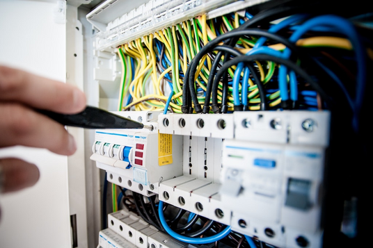 Een close-up van een hand die een gereedschap gebruikt om stroomonderbrekers af te stellen in een elektrisch paneel met meerdere gekleurde draden aangesloten op klemmen.