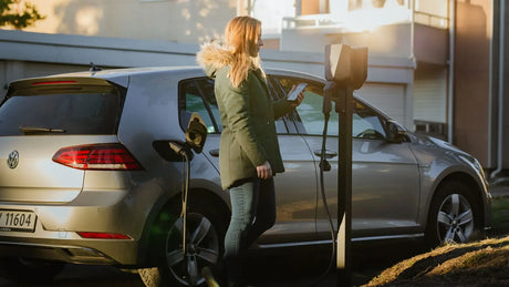 Een vrouw in een groene jas laadt haar zilveren Volkswagen elektrische auto op bij een buitenstation met de Easee Base montagepaal voor Easee laadpunten op een zonnige dag.