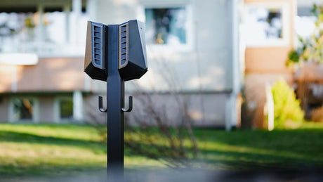 Een zwart dubbel stopcontact laadstation met haken is gemonteerd op een Easee Base montagepaal voor Easee laadpunten, dat buiten op gras staat met een vaag flatgebouw op de achtergrond.