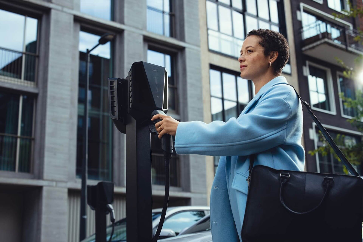 Een vrouw in een lichtblauwe jas staat buiten en houdt een oplaadkabel vast bij een laadpaal voor elektrische voertuigen thuis, met moderne appartementen, geparkeerde auto's en een Easee Charge Max 22kW oplaadpunt elektrisch voertuig op de achtergrond.