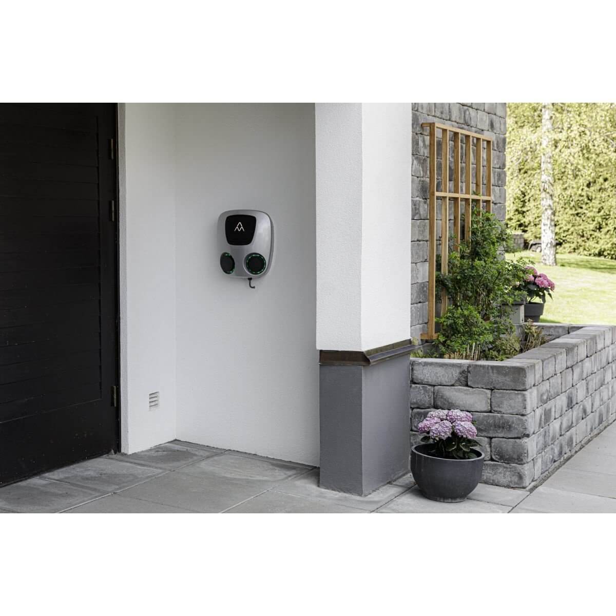 Een Laadstation Charge Amps Aura met Sim 2x22 kW 3-fase 32A bi-directioneel voertuig naar het elektriciteitsnet is gemonteerd op een buitenmuur van het huis in de buurt van een zwarte garagedeur, met bloemen in potten en groen op een stenen veranda op de achtergrond.