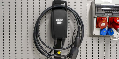 Een zwarte CTEK NJORD GO 3x16A 11kW thuislader met 6,5 meter kabel voor uw elektrische auto is gemonteerd op een geperforeerde grijze muur naast een elektriciteitspaneel met rode en blauwe stopcontacten.