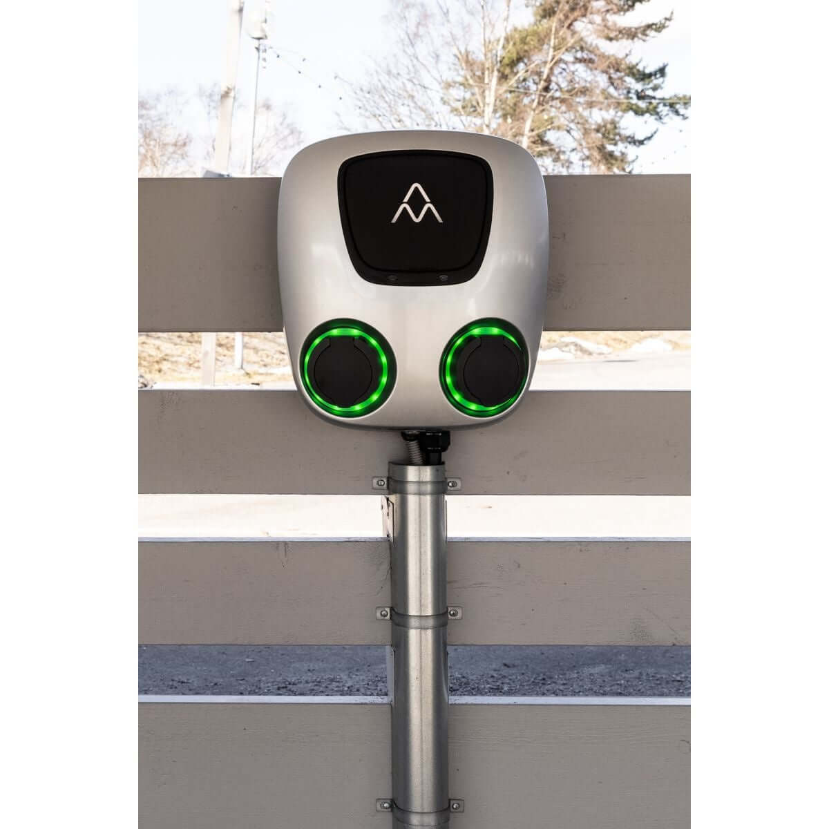 Een Laadstation Charge Amps Aura met Sim 2x22 kW 3-fase 32A bi-directioneel voertuig naar net is gemonteerd op een licht houten hek buiten, ideaal voor elektrische auto laden, met twee groen verlichte poorten, bomen en blauwe lucht op de achtergrond.