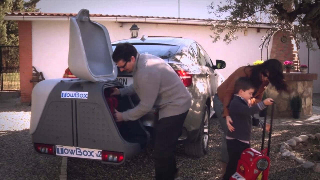 Een man laadt bagage in een Te huur: Bagagebox trekhaakbox Trekhaakbox V2 390 liter achterop een zilverkleurige elektrische auto, terwijl een vrouw voor een huis een jongen helpt met een rode koffer.