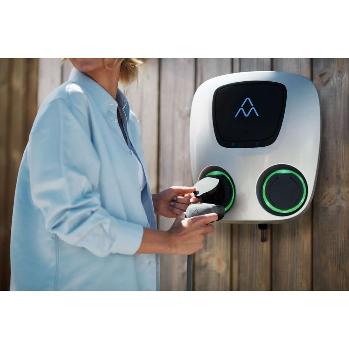 Een persoon in een lichtblauw shirt sluit een laadkabel aan op een Laadstation Charge Amps Aura met Sim 2x22 kW 3-fase 32A bi-directioneel voertuig naar net, met oplichtende groene cirkels, gemonteerd op een houten muur, om een elektrische auto op te laden.