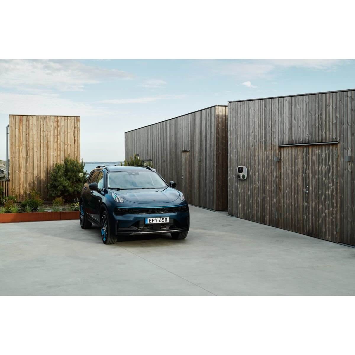 Een blauwe elektrische SUV staat geparkeerd bij moderne houten gebouwen, waar een Laadstation Charge Amps Aura met Sim 2x22 kW 3-fase 32A bi-directioneel voertuig naar net is gemonteerd om op te laden. Links verschijnt groen onder een gedeeltelijk bewolkte hemel.