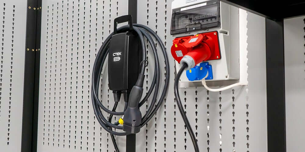 Een zwarte CTEK NJORD¨ GO WALL MOUNT met opgerolde kabel wordt in de houder geplaatst, bevestigd aan een geperforeerde metalen muur naast een stopcontactdoos met rode en blauwe stopcontacten.