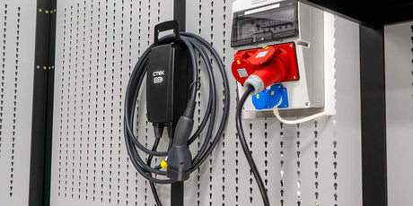 Een zwarte CTEK NJORD¨ GO WALL MOUNT met opgerolde kabel wordt in de houder geplaatst, bevestigd aan een geperforeerde metalen muur naast een stopcontactdoos met rode en blauwe stopcontacten.