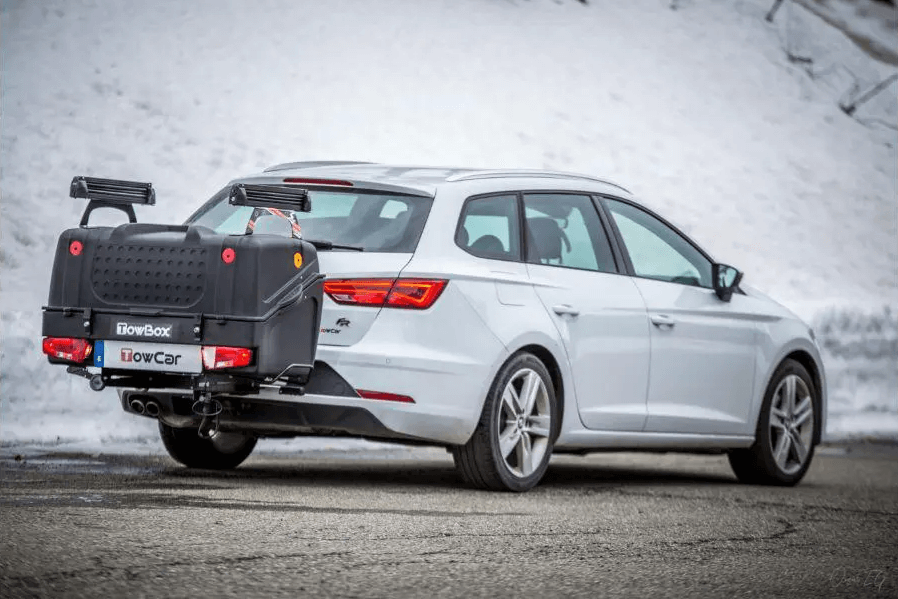 Een witte elektrische stationwagon staat geparkeerd in de sneeuw met een zwarte "Te huur: Towbox V1 met Skidragers - Ook ideaal voor Wintersport" op de trekhaak.