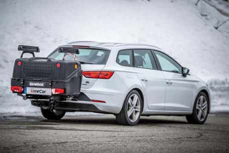 Een witte elektrische stationwagon staat geparkeerd in de sneeuw met een zwarte "Te huur: Towbox V1 met Skidragers - Ook ideaal voor Wintersport" op de trekhaak.
