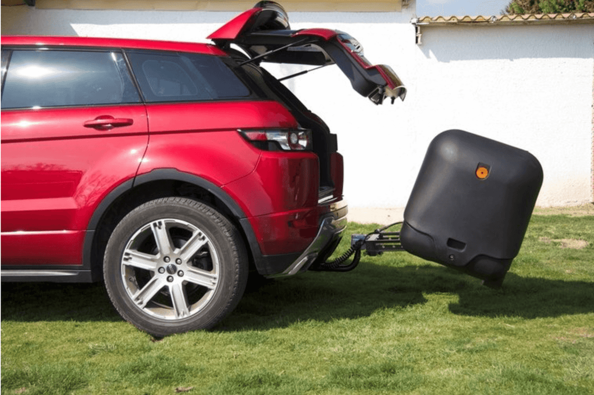 Een rode SUV met geopende achterklep staat geparkeerd op gras en trekt een zwarte Te huur: Bagagebox trekhaakbox Trekhaakbox V2 390 liter aan de trekhaak. Een witte muur en een deel van een gebouw zijn zichtbaar op de achtergrond.