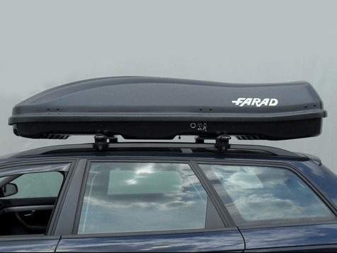 Een Towbox V2 390 liter bagagebox is gemonteerd op een donkerblauwe elektrische auto, die geparkeerd staat onder een bewolkte hemel.