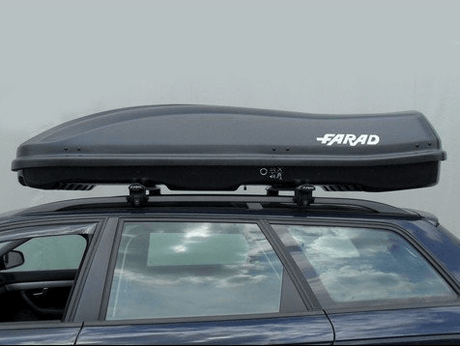 Een Towbox V2 390 liter bagagebox is gemonteerd op een donkerblauwe elektrische auto, die geparkeerd staat onder een bewolkte hemel.