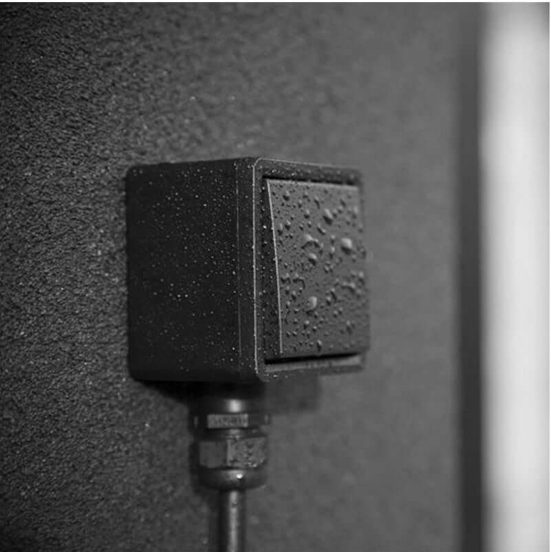 Een close-up van de Wandcontactdoos voor veilig laden met 16A thuislader, een zwarte rechthoekige contactdoos met waterdruppels op het oppervlak, gemonteerd op een donkere muur met structuur - perfect om EV's veilig buiten op te laden.