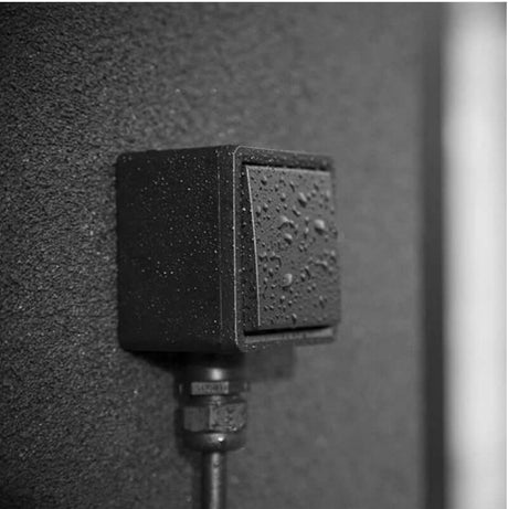 Een close-up van de Wandcontactdoos voor veilig laden met 16A thuislader, een zwarte rechthoekige contactdoos met waterdruppels op het oppervlak, gemonteerd op een donkere muur met structuur - perfect om EV's veilig buiten op te laden.