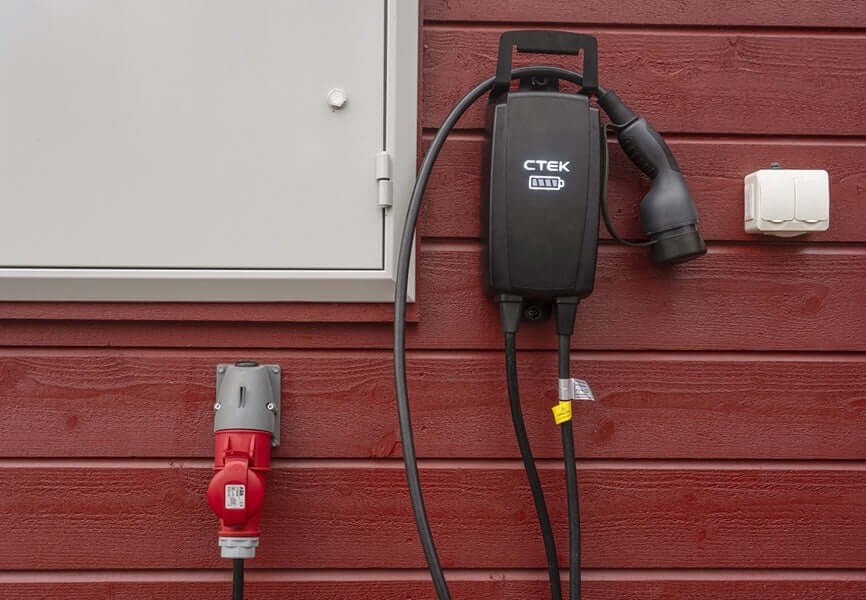 Een zwarte CTEK NJORD GO 3x16A 11kW thuislader met 6,5 meter kabel is gemonteerd op een rode houten wand naast een witte meterkast en een rood stopcontact. De laadkabels hangen netjes omlaag vanaf het toestel.