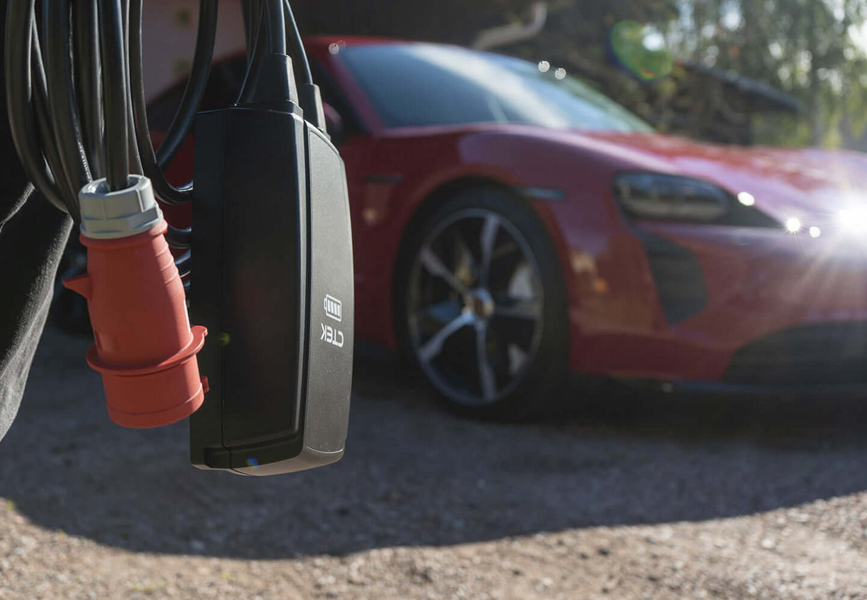 Close-up van een CTEK NJORD GO 3x16A 11kW thuislader met 6,5 meter lange kabeloplaadaansluiting in focus, terwijl een rode sportwagen wazig op de achtergrond geparkeerd staat en het zonlicht van rechts schijnt.