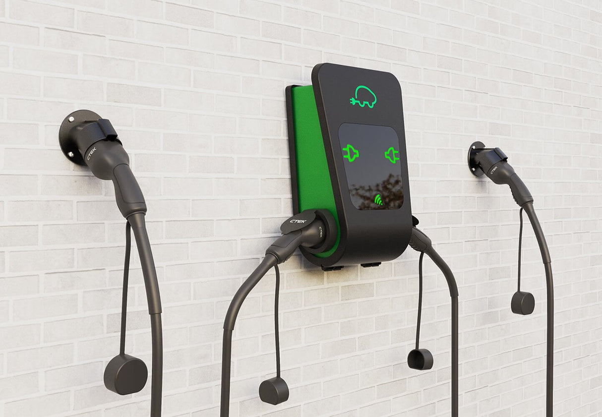 De CTEK Chargestorm Connected 2 11kW/22kW laadpaal voor elektrische auto's wordt aan de muur bevestigd met load balancing, drie laadkabels en stekkers, en een middenunit met groene pictogrammen en een autosymbool op een witte bakstenen muur.