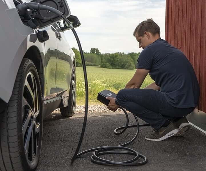 Een persoon hurkt bij een witte elektrische auto die geparkeerd staat op een oprit, en sluit een CTEK NJORD GO 3x16A 11kW thuislader met 6,5 meter kabel aan naast een rood houten gebouw, met groene velden en bomen op de achtergrond.