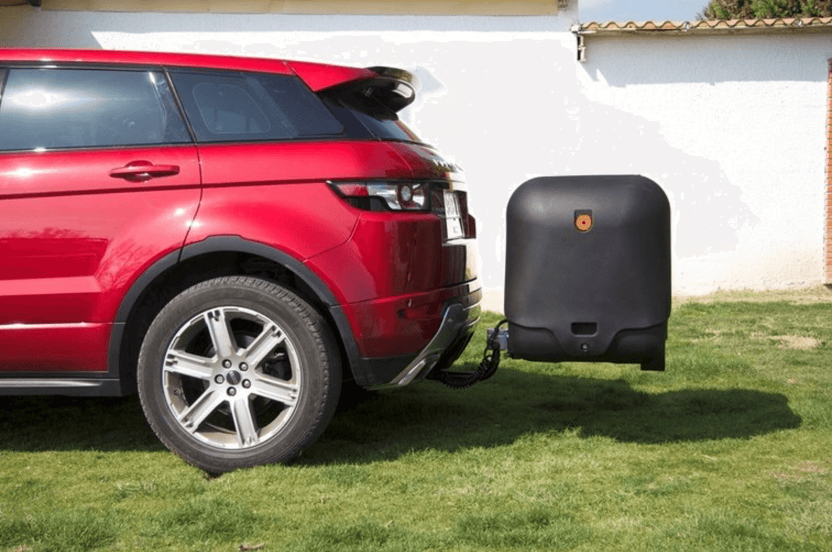 Een rode SUV staat geparkeerd op gras met een "Te huur: Bagagebox trekhaakbox Towbox V2 390 liter" bevestigd aan de trekhaak. Op de achtergrond zie je een witte muur en een deel van een pannendak.