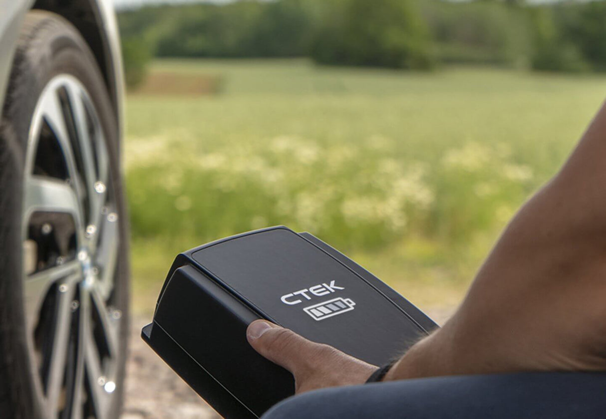 Een persoon houdt een CTEK NJORD GO 3x16A 11kW thuislader met 6,5 meter kabel bij een autowiel, met een groen veld en bomen op de achtergrond, om het gemak voor uw elektrische auto te laten zien.
