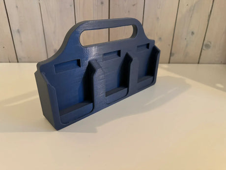 Een zwarte, 3D-geprinte Makita Accuhouder & Transportkar met drie compartimenten en een handvat staat op een wit oppervlak. Licht houten lambrisering vormt de achtergrond.