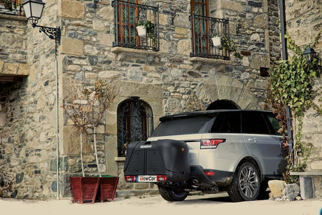 Een zilveren SUV, mogelijk elektrisch, staat geparkeerd bij een rustiek stenen gebouw met potplanten en balkons, met een grote Te huur: Bagagebox Trekhaakbox Trekhaakbox V2 390 liter achterop.