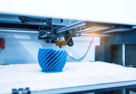 Een 3D printer maakt een blauw object met een spiraalpatroon op zijn printplatform, met de spuitmond van de printer in beweging boven het gedeeltelijk voltooide ontwerp.